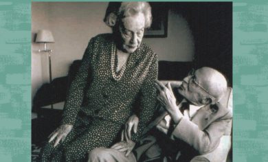 An elderly man is gesturing while seated, speaking to an elderly woman standing next to him.