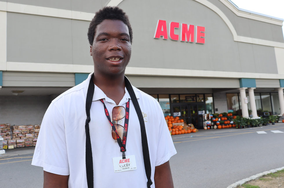 Image of a young person smiling stands in front of the entrance of ACME Supermarket. 