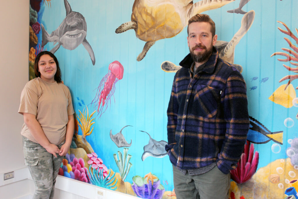 Teacher stands with a student next to the underwater art mural they created.